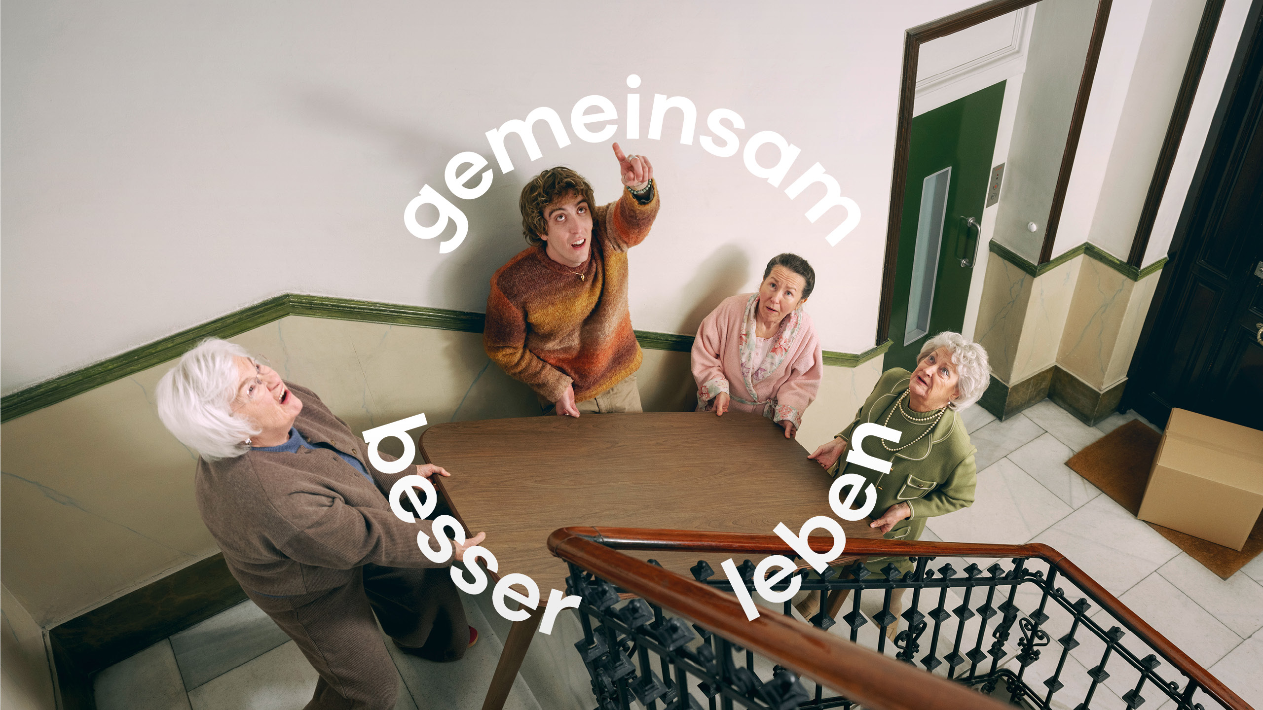 Einem Jungen Mann wird von zwei alten Damen dabei geholfen einen Tisch das Stiegenhaus hochzutragen.